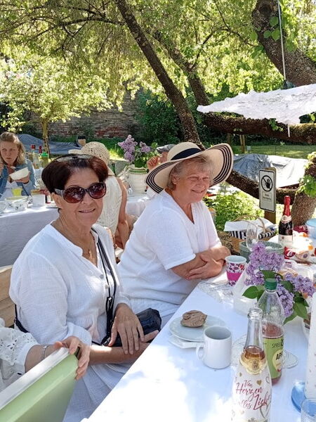 2 Frauen in weißer Kleidung, die linke mit großer Sonnenbrille, die rechte mit großen Sonnenhut sitzen an einem Picknicktisch und lächeln.
