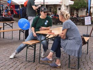 Zwei Frauen sitzen an einer Biertischgarnitur und unterhalten sich. Frau mit Base Cape, Sonnenbrille, hält einen SEKIZ Luftballon in der Hand. Die Frau die ihr gegenübersitzt ist blond, mit blau-weißem Kleid.