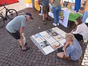 Vor einem Stand. Alter Mann beugt sich nach vorn um eine Art Collage zu betrachten. Bei der Collage sitzt ein Mann auf dem Boden, eine Frau mit langen brauen Haaren hockt daneben.