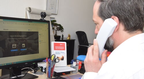 Mann an einem Schreibtisch am PC mit Telefonhörer am Ohr und Stift in der anderen Hand. Mann hat graue Haare und Bart, er trägt ein weißes langärmliches Hemd.