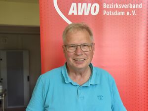 Portrait. Älterer Mann mit Brille und grauen Haaren. Er trägt ein blaues Poloshirt. Er steht vor einem roten AWO RollUp Banner.