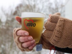 Teetasse mit AWO-Logo. Tasse wird mit zwei Händen in fingerlosen braunen Handschuhen gehalten.