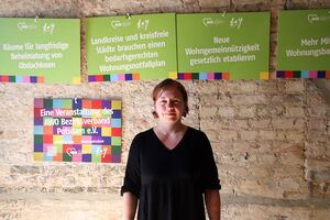 Portrait. Junge Frau. Kurze braune Haare. Sie trägt ein schwarzes Shirt mit V-Ausschnitt. Sie steht vor einer Steinwand. An der Wand sind Schilder mit Forderungen: Landkreise und kreisfreie Städte brauchen einen bedarfsgerechten Wohnungsnotfallplan