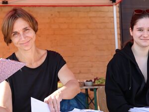 Zwei Frauen sitzen draußen. Eine im schwarzen T-Shirt, mit brauen Haaren. Daneben, eine Jugendliche mit Sonnenbrille auf den Kopf geschoben und schwarzer Kapuzenjacke. Beide lächeln in die Kamera.