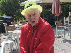 Seniorin mit Rollator auf sonniger Terrasse. Sie trägt rote Kleidung und einen gelben Hut.