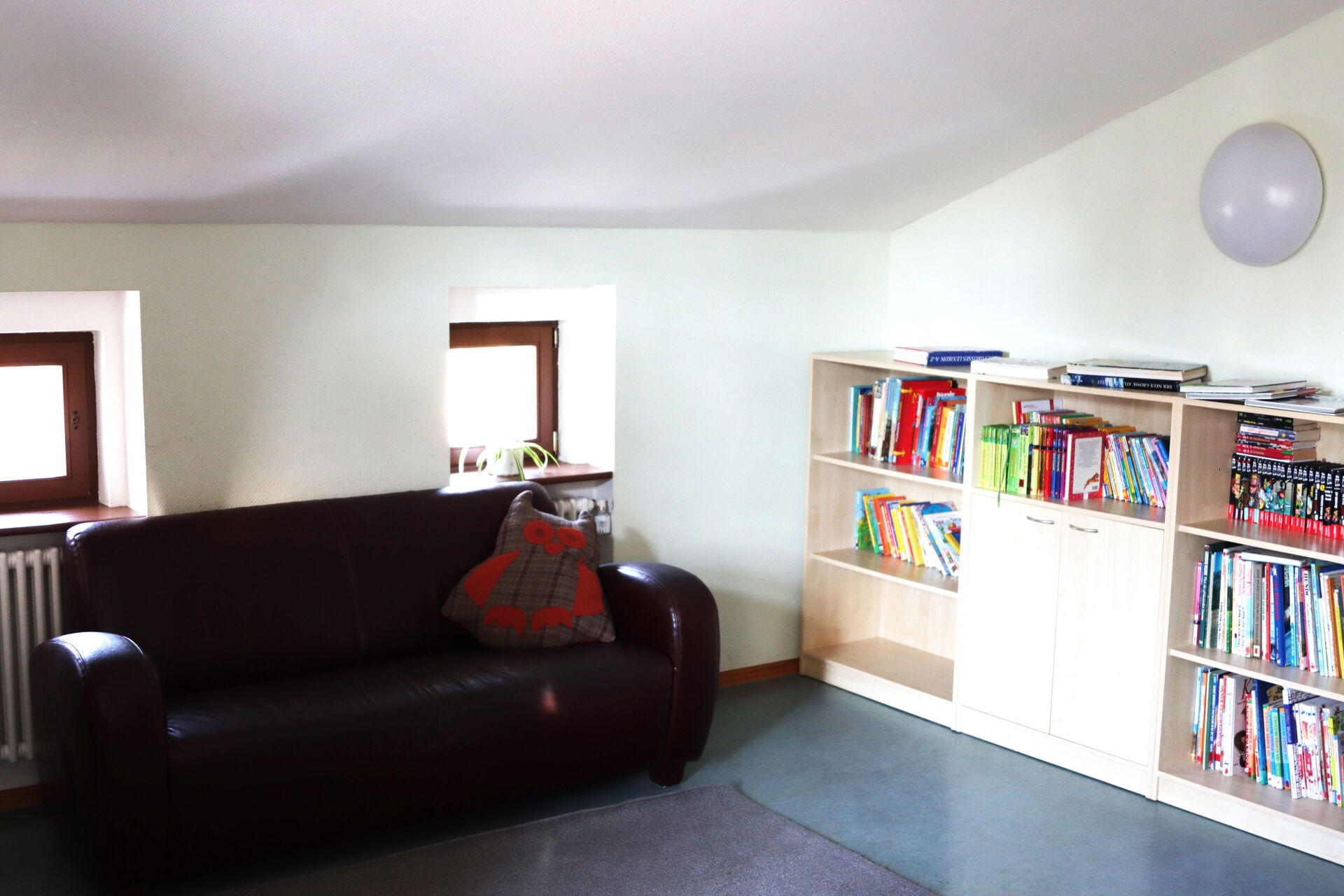 Aufenthaltsraum. Dachgeschoss mit Dachschräge, kleine Fenster. Eine schwarze Couch vor den Fenstern. Regal an der Wand stehend mit Büchern.