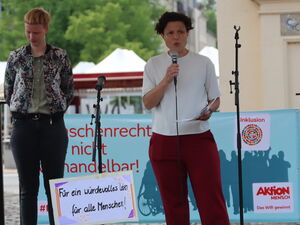 Eine Bühne mit Gebärdendolmetschung und einer Frau mit Mikrofon. Dabor Protestplakate: Demokratie braucht Inklusion.