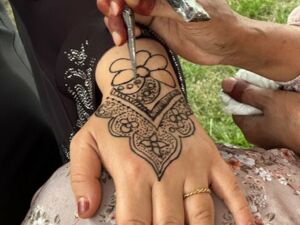 Frauen Hand mit roten Fingernägeln und Ring am Ringfinger. Die Hand liegt auf einem hellen Tuch mit Blumenmuster und es wird ein kunstvolles Henna Tattoo darauf gemalt.