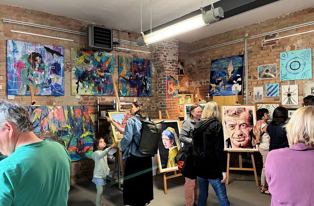 Menschen in einem Atelier, die sich Kunst Bilder, Gemälde ansehen. Es sind abstrakte Bilder aber auch Portraits darunter. Eine Frau im gelben Gewand und das Gesicht eines älteren Mannes mit Falten.