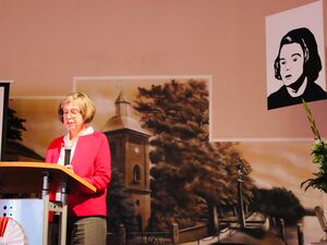 Frau am Rednerpult. Frau mit Brille, brauen Haaren, weißer Bluse und rotem Blazer darüber. An der Wand ein Schwarz-Weiß Bild von Sophie Scholl