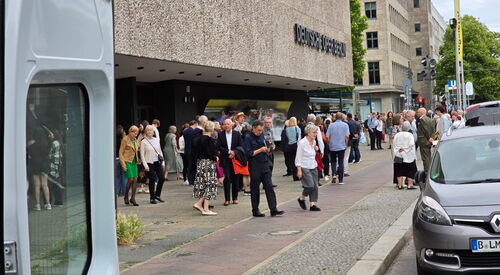Auf einem Gehweg an einer Straße vor der Deutschen Oper Berlin. Viele Menschen stehen vor der Oper.