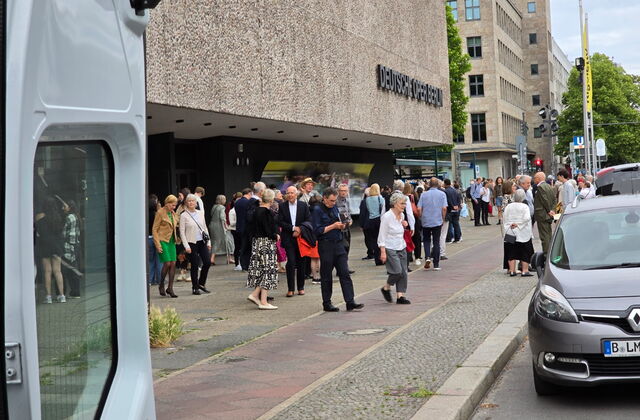Auf einem Gehweg an einer Straße vor der Deutschen Oper Berlin. Viele Menschen stehen vor der Oper.