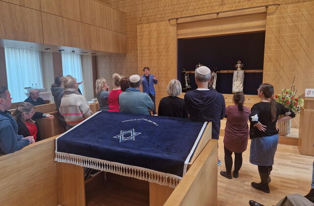 Gruppe steht verteilt im Synagogen Raum und hört dem Mann mit dem blauen Sakko zu.