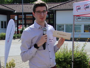 Im Freien. Junger Mann mit Mikrofon in der rechten Hand und einem Holzklotz in der Linken Hand. Auf dem Holzklotz steht das Wort: Frieden. Mann hat kurze dunkle Haare, Brille und trägt ein helles Hemd.