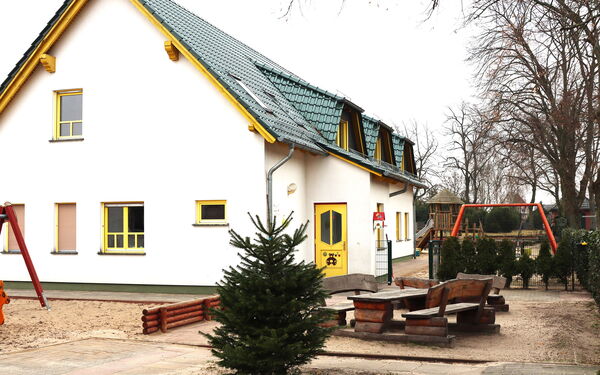 Weißes Haus mit gelber Tür und gelben Fensterrahmen, das Dach ist grün. Kahle Bäume, zwei Holzbänke mit Holztisch, ein kleiner Tannenbaum und Spielgeräte ums Haus verteil. Schaukel, Klettergerüst mit Rutsche.
