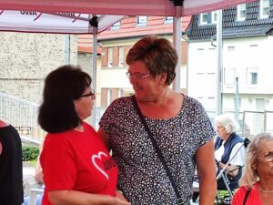 Zwei Frauen die sich in die Augen blicken und sichh unterhalten. Eine Frau im roten AWO T-Shirt mit schwarzen vollen Haaren und Brille und eine füllige Frau mit Kurzhaarfrisur, Brille und weißgetupften T-Shirt, lächelt..