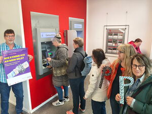 Menschen-Schlange vor Geldautomat. Einige Frauen halten ein großes P In die Kamera. Ein Mann steht links außen und hält ein lila Plakat "Wie komme ich an mein Geld?"