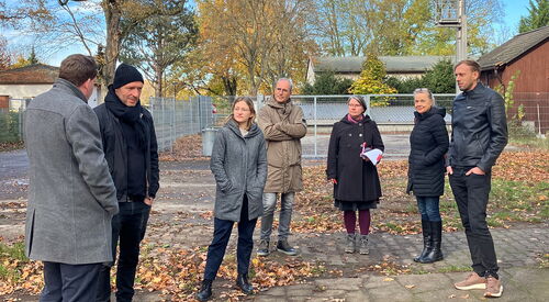 Herbst, Draußen, blauer Himmel, Schäfchenwolken. 7 Personen stehen draußen verteilt. Alle sehen zu Brain Nickholz.