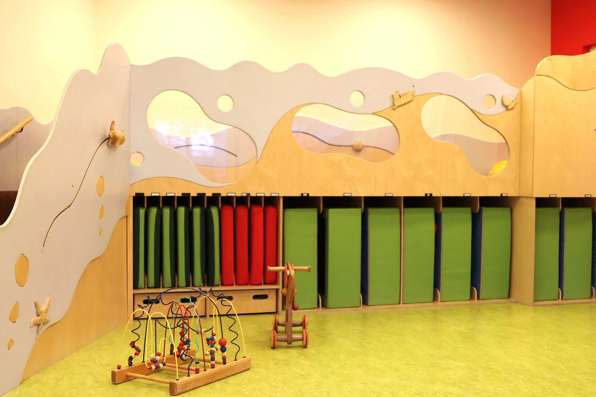 Teil eines hellen Raumes, der wie ein Spielbereich für Kinder gestaltet ist. Dominierend ist eine große, freistehende Holzeinrichtung, die an eine Wellenlandschaft erinnert.