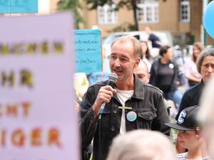 Mann mit Mikrofon, schwarzer Jeansjacke, drunter weißes T-Shirt.Er spricht zu den umstehenden Prostetierenden, die ihre Prodestschilder hoch halten.