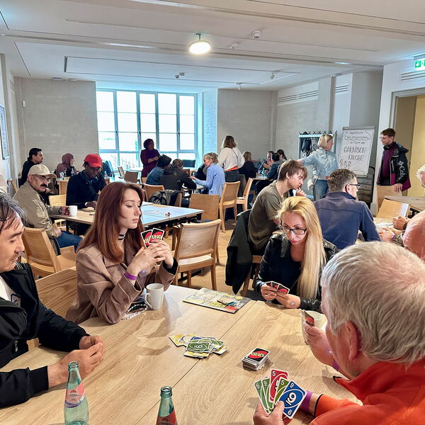 Menschen sitzen in Gruppen an Tischen, am Tisch im Vordergrung wird UNO gespielt.