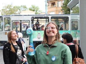 Junge Frau mit schulterlangen braunen Haaren und Brille, sie trägt einen grünen Pullover macht Seifenblasen mit einer Seifenblasenmaschine. Sie steht an einer Tram Haltestelle. Hinter ihr steht eine alte Tatra Straßenbahn.