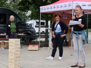 Draußen. Ein Jenga-Turm steht auf dem Boden. Im Hintergrund ein AWO Pavillon. 3 Frauen stehen herum. Blonde Frau mit Brille auf den Kopf geschoben, schwarzem dünnen Pulli, hellgrauer Hose, hält Mikro in der Hand, Notizen in der anderen Hand und spricht.