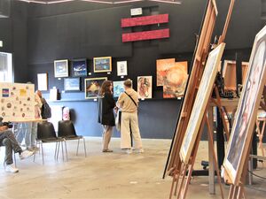 Galerieraum. Bilder hängen an der Wand. Auf Staffeleien sind weitere Bilder im Raum ausgestellt. Es gibt ein paar Stühle, ein Mann sitzt und betrachter die bilder. Zwei Menschen stehen vor den Bilder an der Wand.