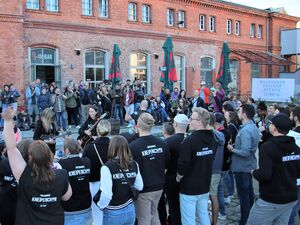 Viele Menschen vor einem rotenn Backsteingebäude. In der Mitte der Menschen sind Musikerinnen mit Gitarren. Viele Leute tragen Kleidung mit Aufdruck vom Potsdamer Kneipenchor.