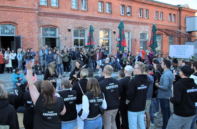 Viele Menschen vor einem rotenn Backsteingebäude. In der Mitte der Menschen sind Musikerinnen mit Gitarren. Viele Leute tragen Kleidung mit Aufdruck vom Potsdamer Kneipenchor.