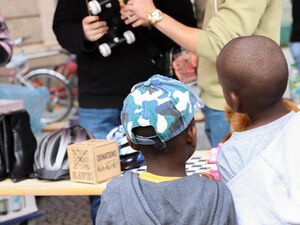 Zwei kleine schwarze Kinder stehen vor einer Holzbank auf der unterschiedliche Dinge stehen / liegen. Schwarze Damenschuhe, zwei Fahrradhelme und eine Donations Wanted Würfel Spardose.