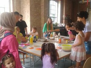 Lichtdurchfluteter Raum durch große Fenster. Kinder und Erwachsene unterschiedlicher Nationen an einem Tisch. Farbenschleuder und Farben stehen auf dem Tisch.