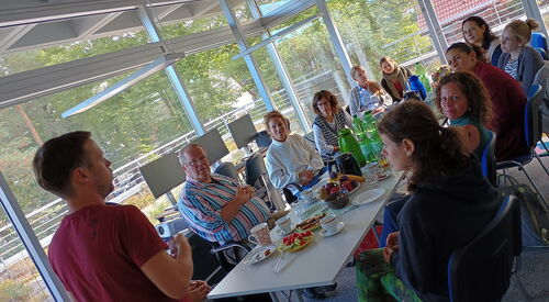 Verglaaster Konferenzraum. 10 Menschen sitzen an einem gedeckten Tisch. An der Stirnseite steht ein Mann im roten T-Shirt auf dem die Aufmerksamkeit ruht.