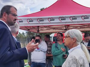 Menschen mit Fotoaperat und Handys stehen unter einem roten AWO Pavillon. Eine Frau mit langen weißen zum Zoüf gebundenen Haaren redet mit einem großen Mann im Anzug und mit Brille.