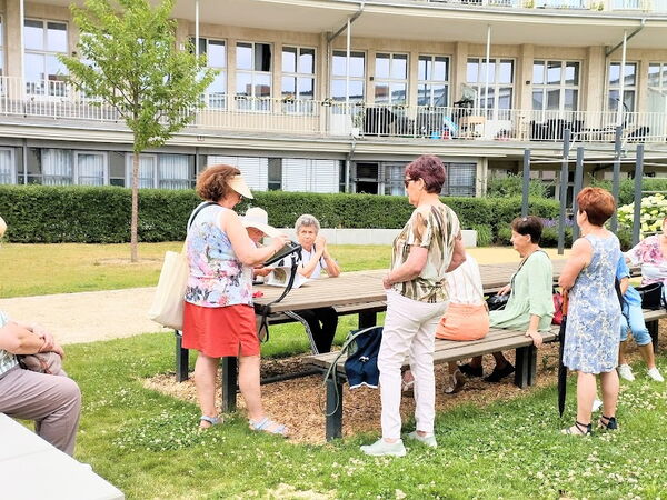 Draußen, schön angelegte Grünanlage vor einem Gebäude mit Balkonen. Einige Frauen sitzen an Picknicktischen.