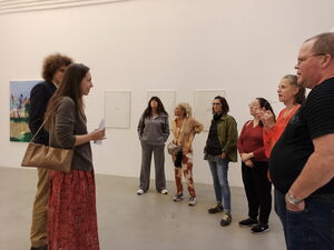 Großer weißer Raum mit Bilderrahmen an der Wand. Frauen und ein fülliger Mann stehen im Halbkreis. Zwei Personen stehen vor ihnen.