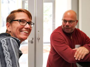 Im Eingangsbereich. Frau lächelt in die Kamera, ein Mann steht an einen Stehtisch gelehnt.