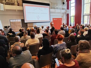 Präsentation an der Leinwand vorn im Raum. Viele Teilnehmende auf Stühlen.