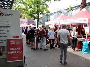 Blick aufs Gelände von Eingang her. Ein AWO Aufsteller mit Text: Herzlich willkommen.     Menschen auf dem Gelände sehen sich um, holen sich Bratwurst und Burger.