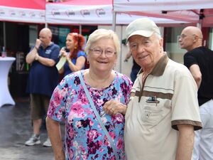 Draußen. Zwei ältere Menschen stehen eng beieinander und lächeln in die Kamera. Frau mit geblümten blauen T-Shirt, sie trägt Brille. Mann mit Basecap und Poloshirt beides in beige.