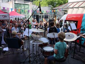 Draußen. Bühne mit Instrumenten und Kindern von Hinten. Blonder Junge am Schlagzeug, weitere Kinder an Keyboard, Mikrofone. Vor der Bühne viele Gäste die sich das Programm ansehen / anhören.