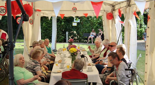 Draußen, Festzelt, mit rot-weißen Wimpel, roten und weißen Luftballons geschmückt. Lange Festtafel an der Senioren mit Kaffee und Kuchen sitzen, Das Wetter ist sonnig.