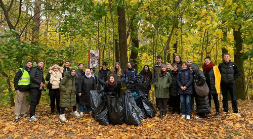 Rund 25 Menschen von Jung bis älter stehen gemeinsam vor einem Wald. Es liegt Laub auf dem Boden aber die Baume haben noch Laub. Vor der Gruppe mehrere gefüllte, schwarze Müllsäcke. Alle blicken stolz und fröhlich zur Kamera.