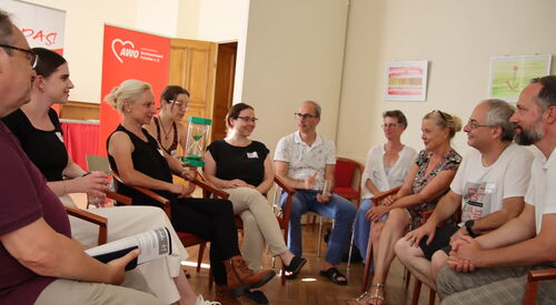 10 Menschen sitzen in einem Raum auf Stühlen. Eine Frau hat eine grüne sanduhr auf dem Schoß, die andere sitzen sich gegenüber.