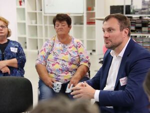 In einem Ladenraum. Zwei Frauen von vorn im äußeren Stuhlkreis. Ein Mann mit kurzen brauen Haaren, Dreitagebart, weißem Hemd und blauer Anzugjacke sitzt im inneren Kreis und diskutiert mit.