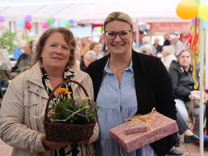 2 Frauen, eine mit Blumenkörbchen,  die andere mit Geschenken. Beide lächeln in die Kamera.