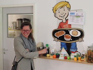 Eine Frau steht lachend an einem kleinen Regal mit gesunden Lebensmitteln. An der Wand ein großes Bild eines blonden Jungen mit Tablett und Speisen darauf.