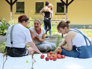 Drei Frauen sitzen auf der Erde. Ein weißes Tuch auf dem Äpfel lieben. Zwei der Frauen sitzen auf dem Tuch mit dem Rücken zur Kamera.