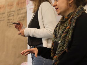 2 Frauen stehen neben einem Flipp Chart mit Notizen, eine hält einen Stift in der Hand und die Andere spricht.