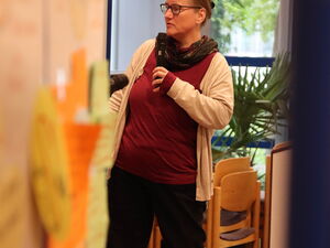 Frau von weiter weg. Sie wirkt konzentriert. Sie steht, den Blick nach links. Sie trägt; Brille, Halstuch, helle Strickjacke über einem roten Oberteil und eine schwarze Hose.
